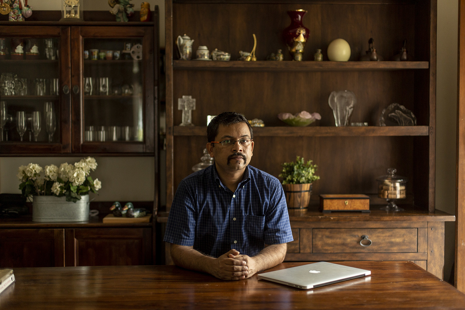 A man sits at a desk, looking into the camera with his hands folded