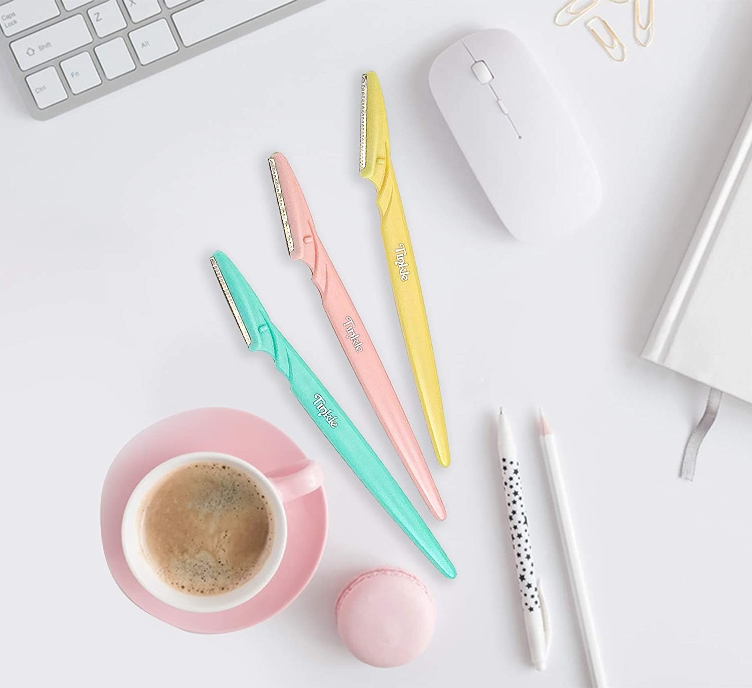 Three eyebrow razors on a desk next to coffee, pencils, a journal, and computer