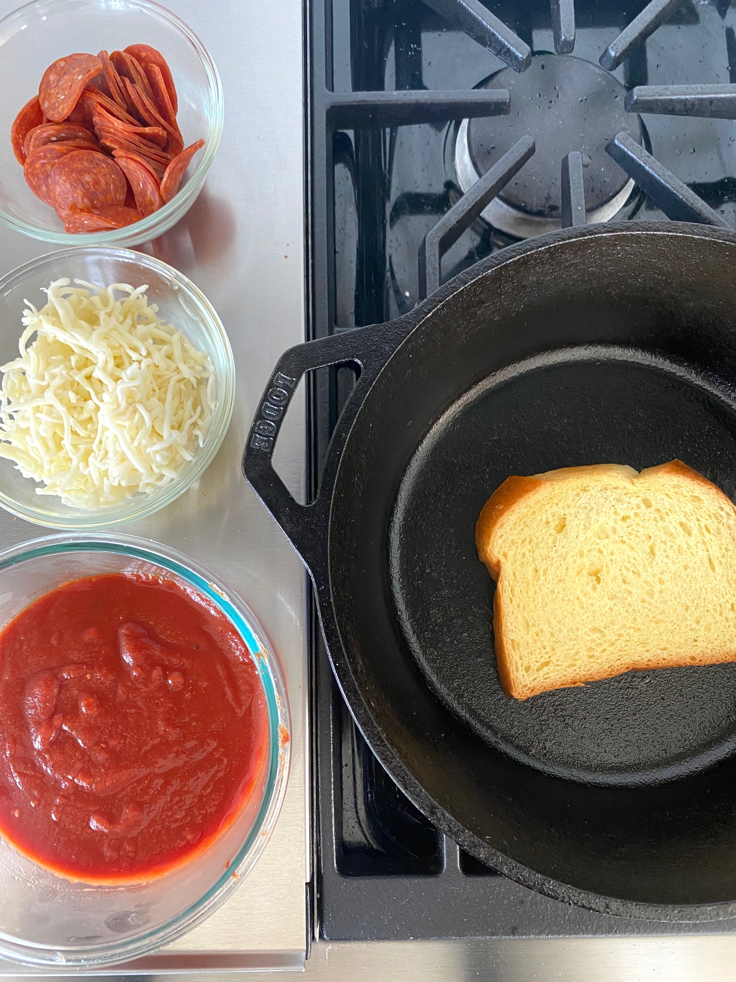 Tomato sauce, cheese, and pepperoni in small bowls next to a cast iron skillet with bread toasting inside.