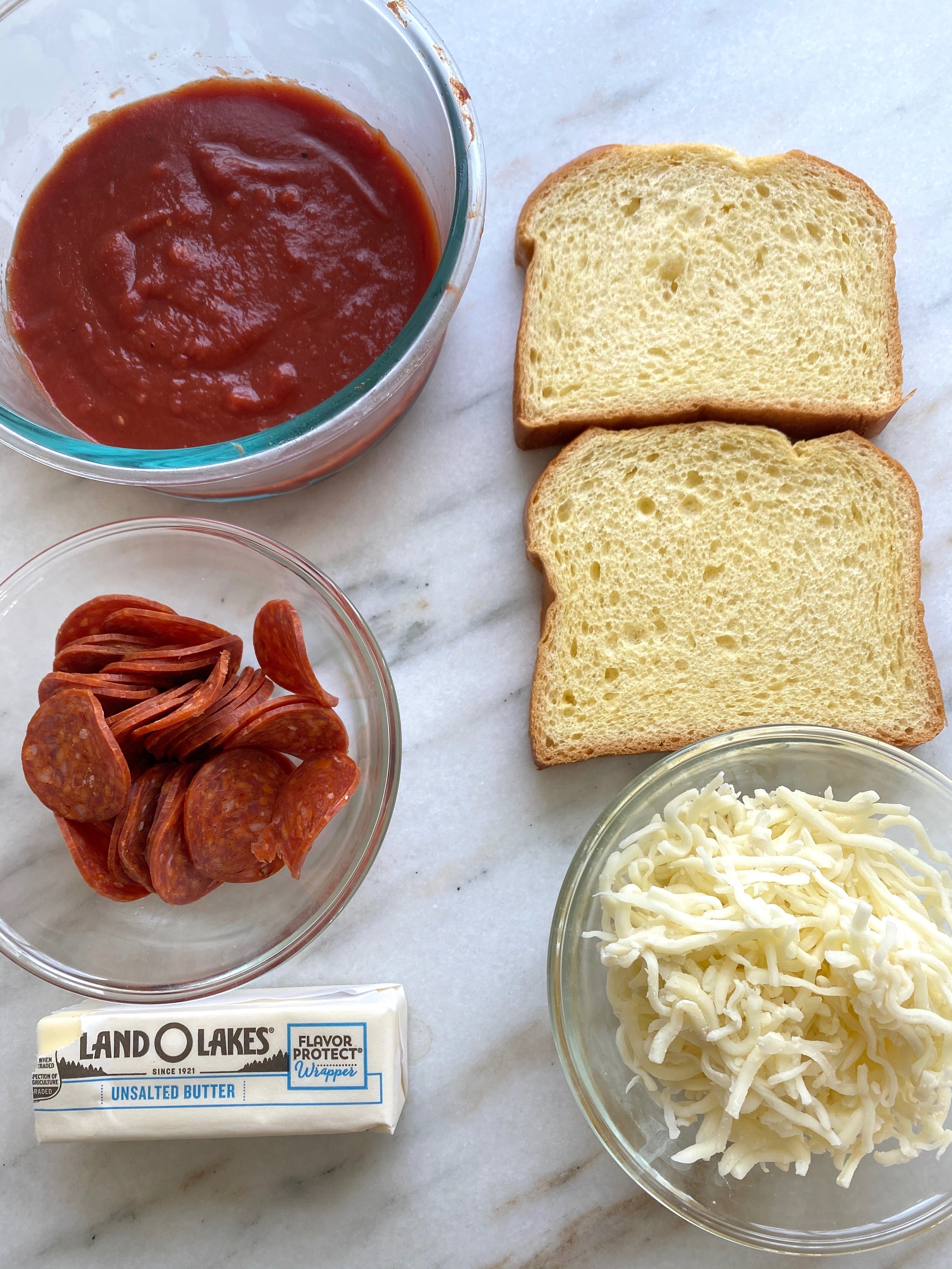 Ingredients for mountain pie on a countertop including shredded mozzarella, butter, pepperoni, tomato sauce, and two slices of brioche bread.