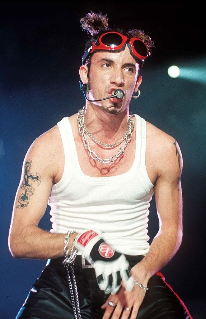 AJ McLean with goggles resting on his forehead as he performs onstage wearing a sleeveless T-shirt, lots of chains, and one glove