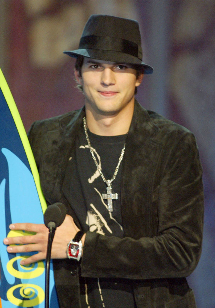 Ashton Kutcher in a stingy brim fedora hat and standing at a microphone and holding a surfing board