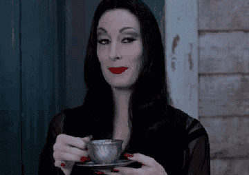 Morticia Addams raises her eyebrows as she brings her teacup to her mouth