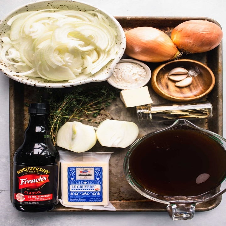 Ingredients for French onion soup on a baking sheet including chopped onions, Gruyère, garlic, herbs, and broth.
