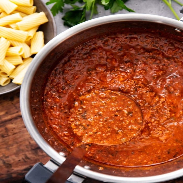 A ladle taking a scoop of Bolognese sauce from an Instant Pot and a bowl of pasta on the side.