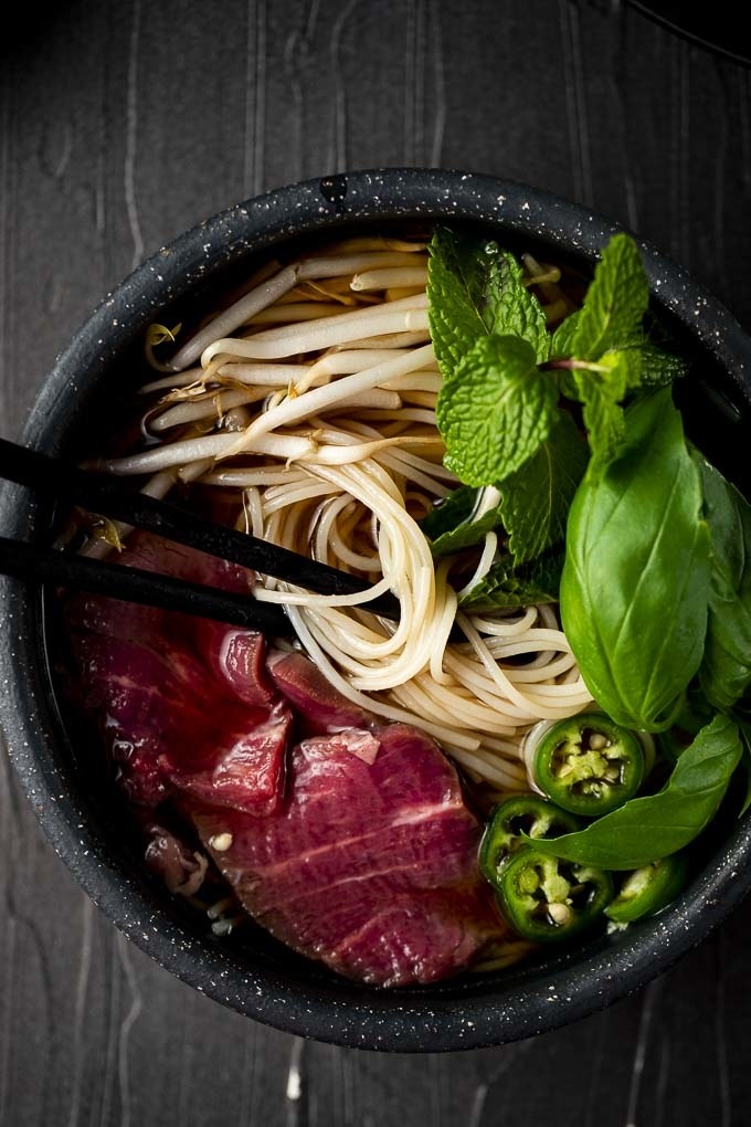 A bowl of pho with noodles, sliced rare beef, jalapeños, and basil.