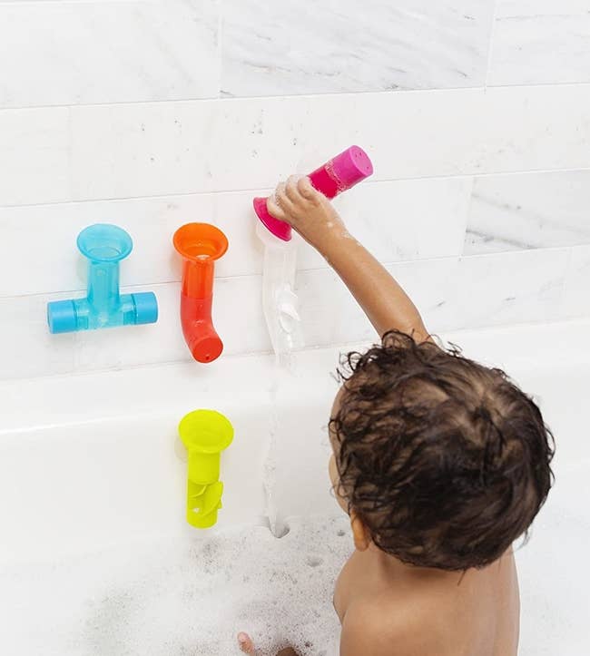 Baby sitting upright and pouring water into pipes attached to bathtub wall 