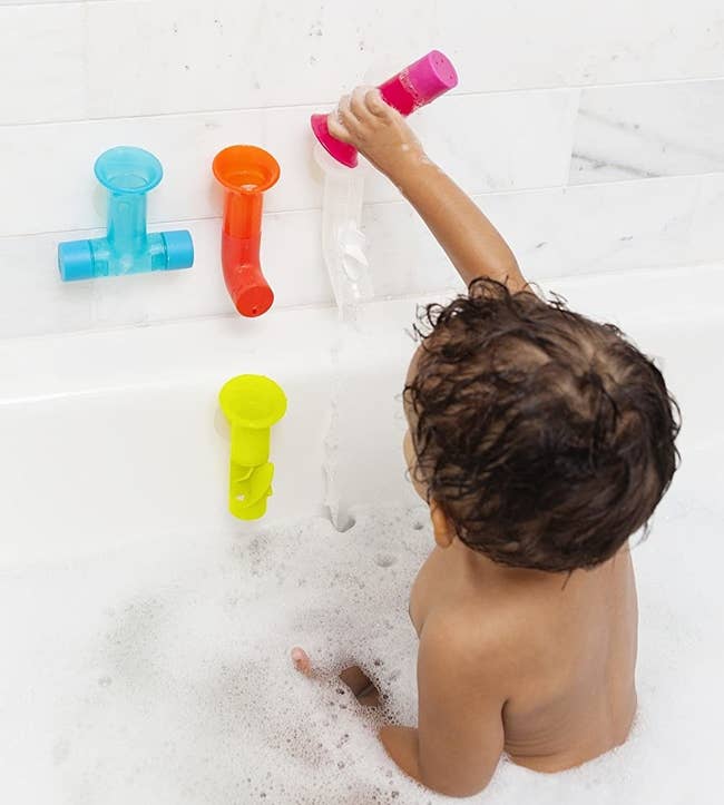 Baby sitting upright and pouring water into pipes attached to bathtub wall 