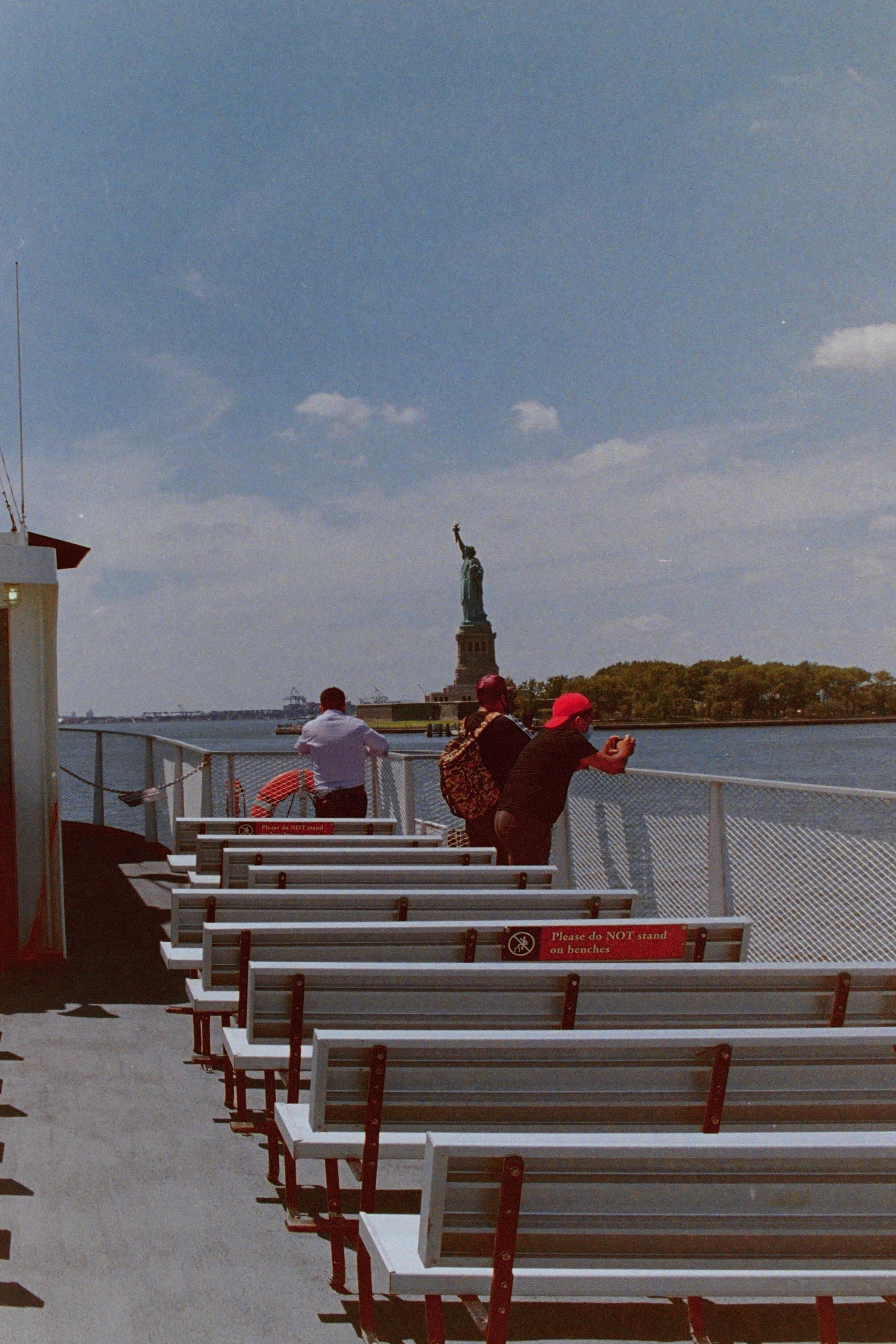 Three people, peering over the water at the Statue of Liberty, stand on a mostly empty ferry