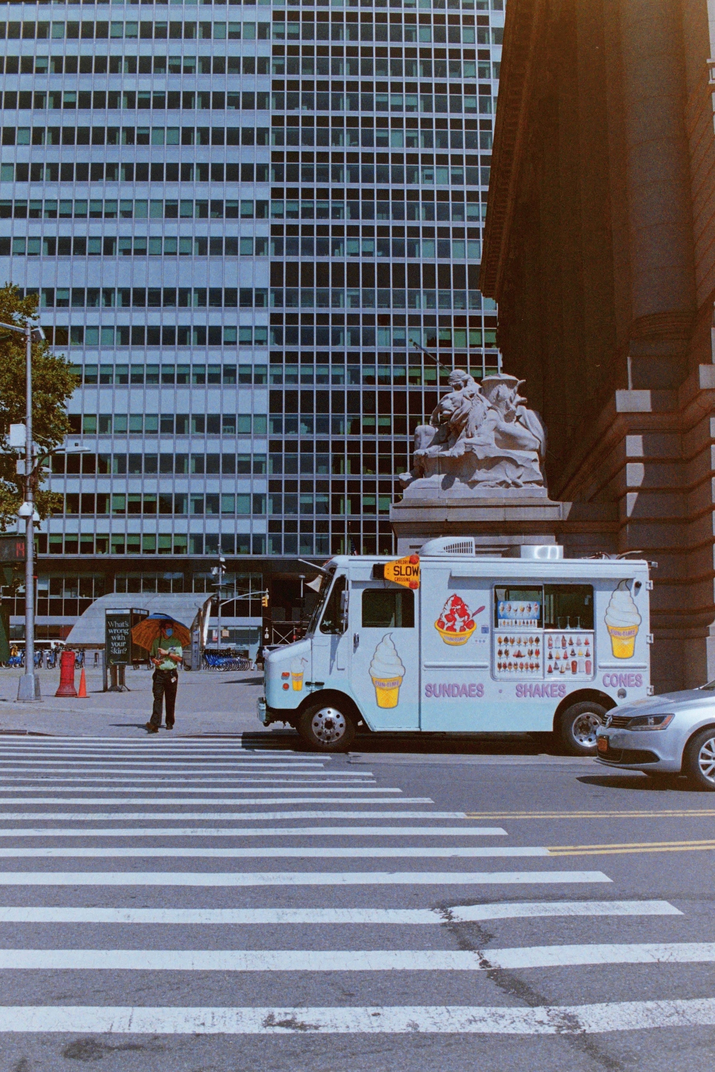 A man, standing under an umbrella and wearing a face mask, looks at an ice cream truck and is about to step into a crosswalk