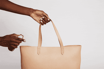 A model hooks the hat holder to the strap of a tote bag, then clamps the hat by its brim between the magnets of the hat holder and lifts the bag