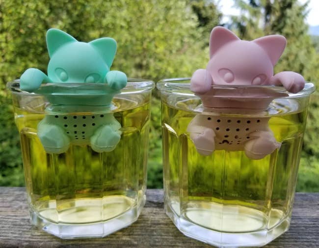 Reviewer's teal and pink tea infusers that look like kittens hanging off the inside edge of mugs with yellow tea inside.  