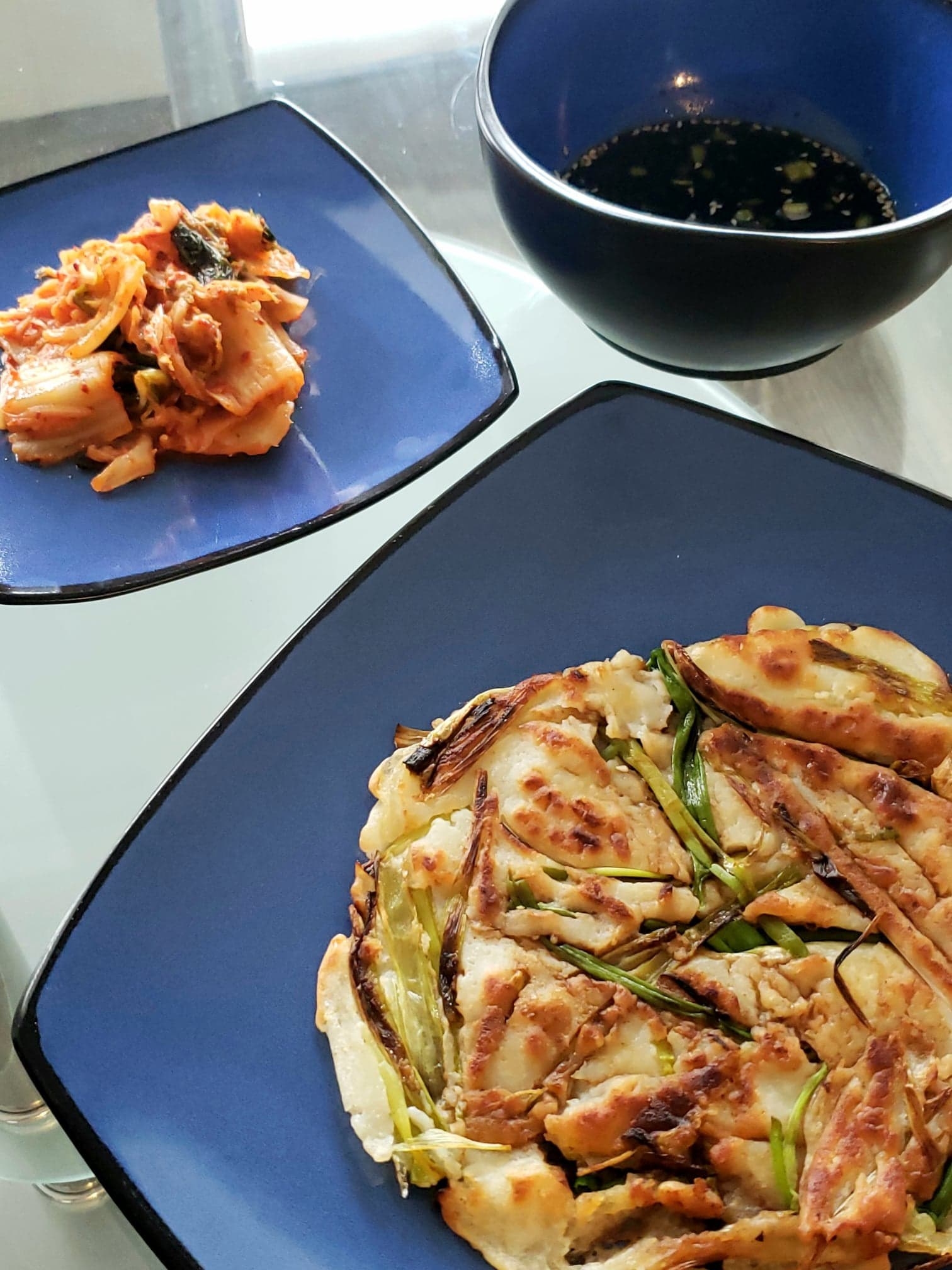 The plated pancake next to a smaller plate of kimchi and the dipping sauce on a table, ready to be eaten