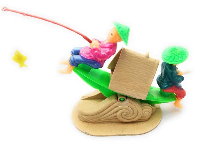 Sea saw ornament in the design of a boat, with two people sitting on either side and facing outwards - one of them holding a fishing rod.