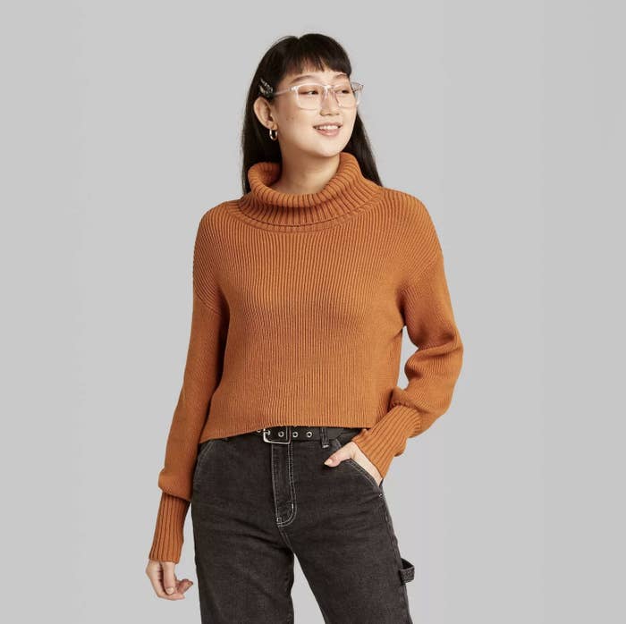 Model is wearing a rust ribbed cropped turtleneck sweater and black jeans.