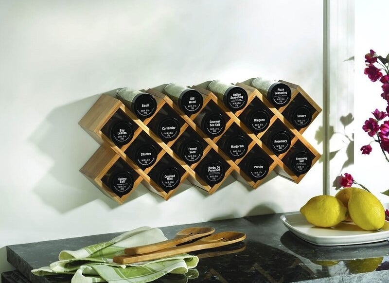 A wooden wall-mounted spice rack holding 18 labeled jars of spices