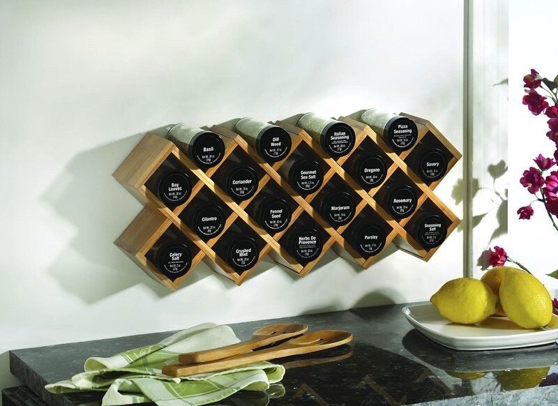 A wooden wall-mounted spice rack holding 18 labeled jars of spices