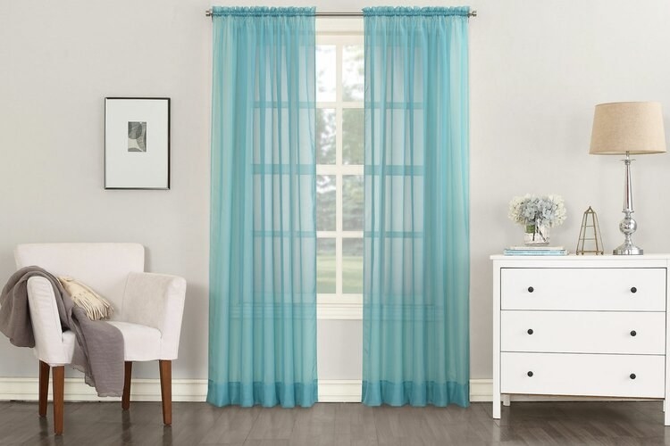 Slightly see-through blue curtains draped from the top of a window to the floor, extending beyond the side edges of the window, and making it the focal point of a neutral-toned room, between a sitting chair and dresser