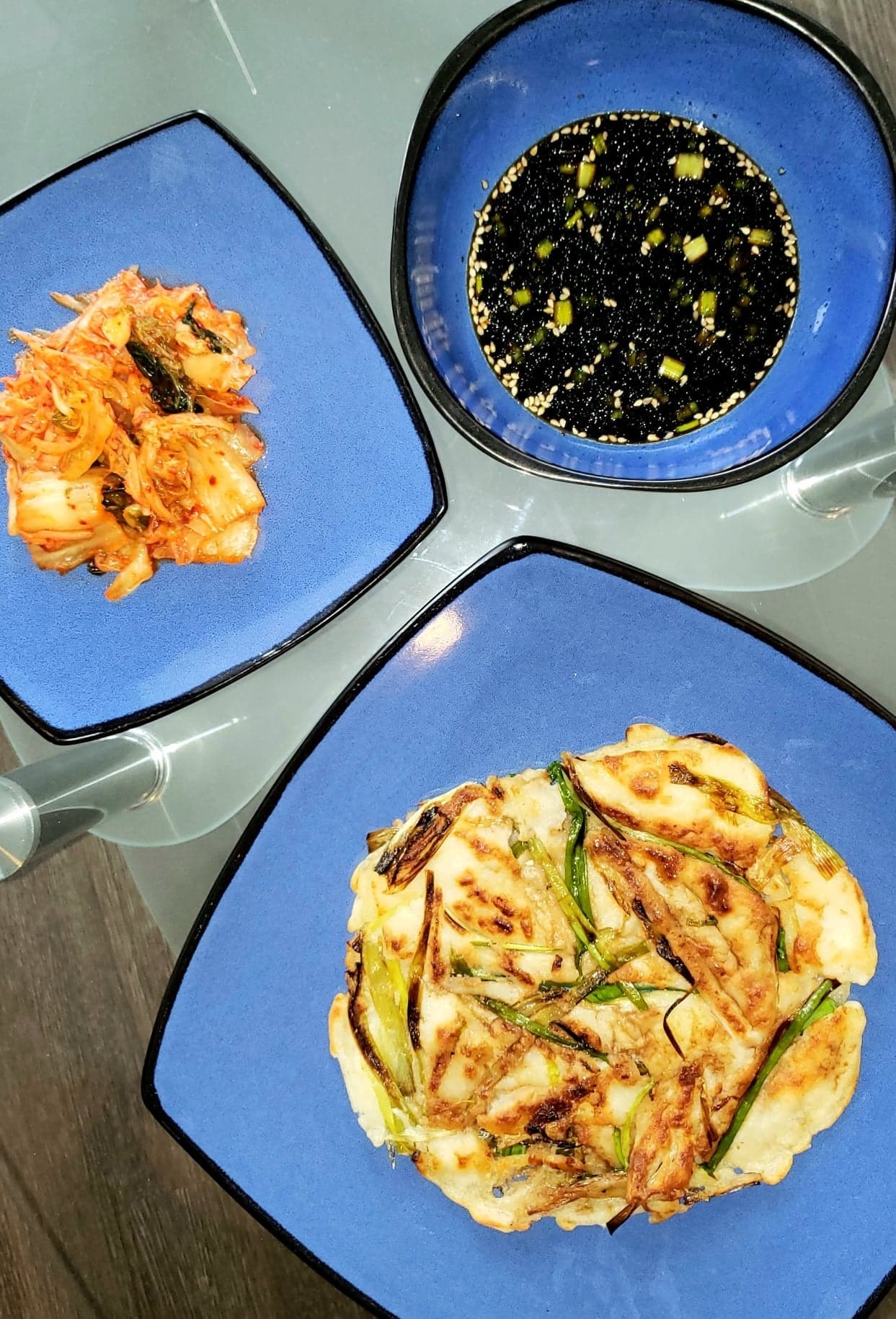 A top down view of the Korean pancake, a plate of kimchi, and the dipping sauce on a table