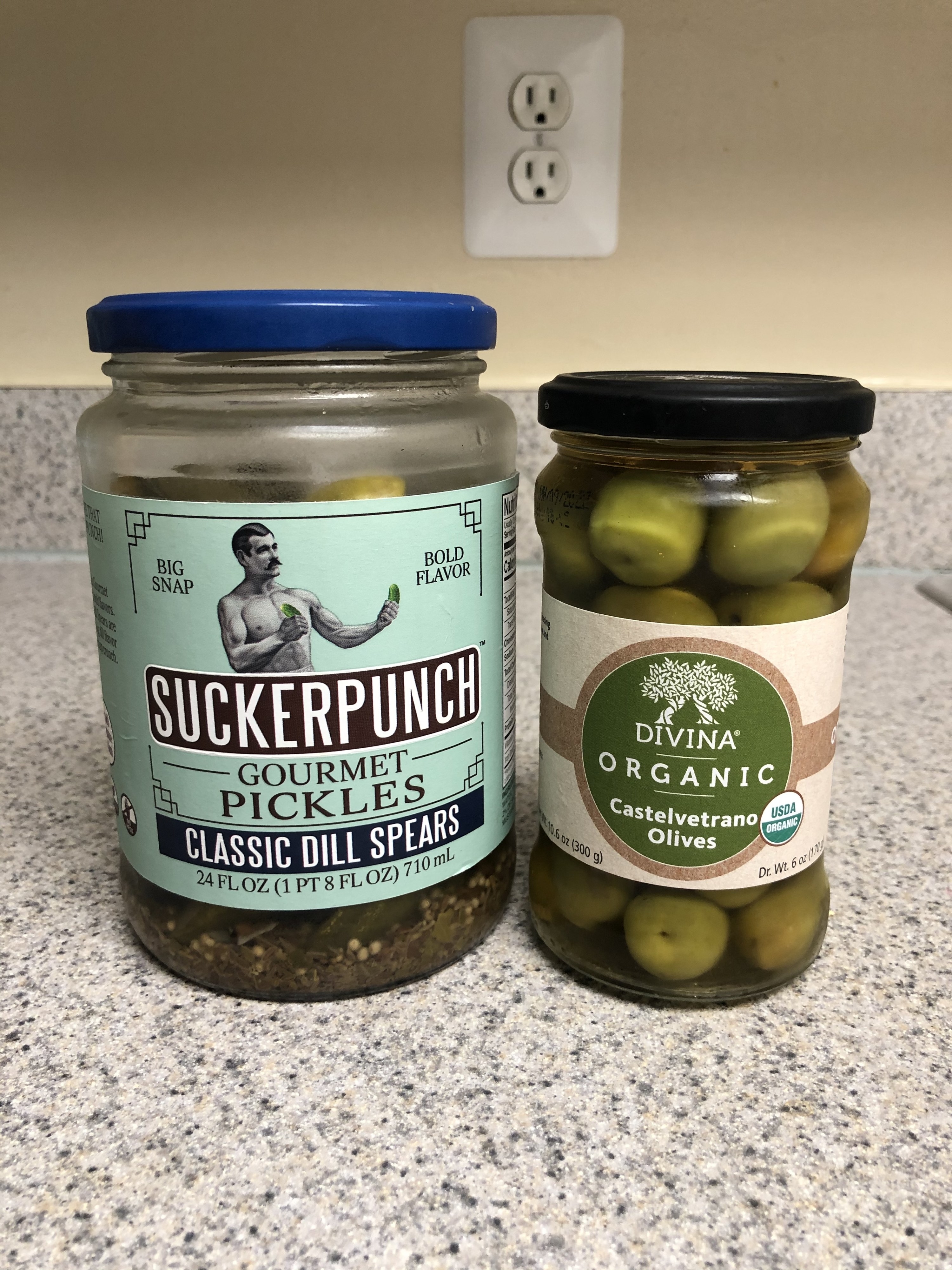 A jar of pickles next to a jar of green olives.