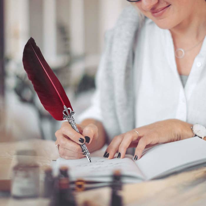 A woman writing on a notebook with a red-feathered quill pen.