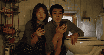A woman and a man sit close together next to a toilet while scrolling on their phones