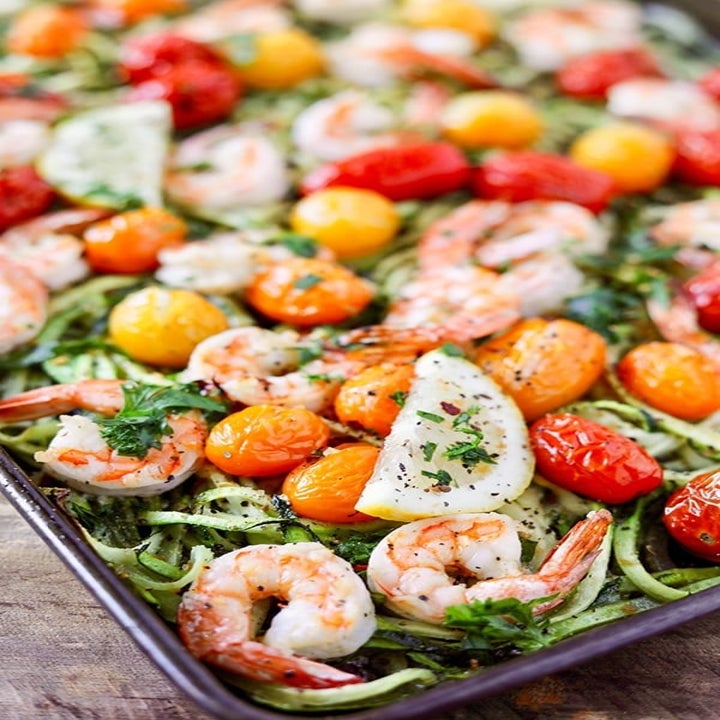Shrimp, zucchini noodles, blistered tomatoes, and lemon with fresh herbs on a sheet pan.