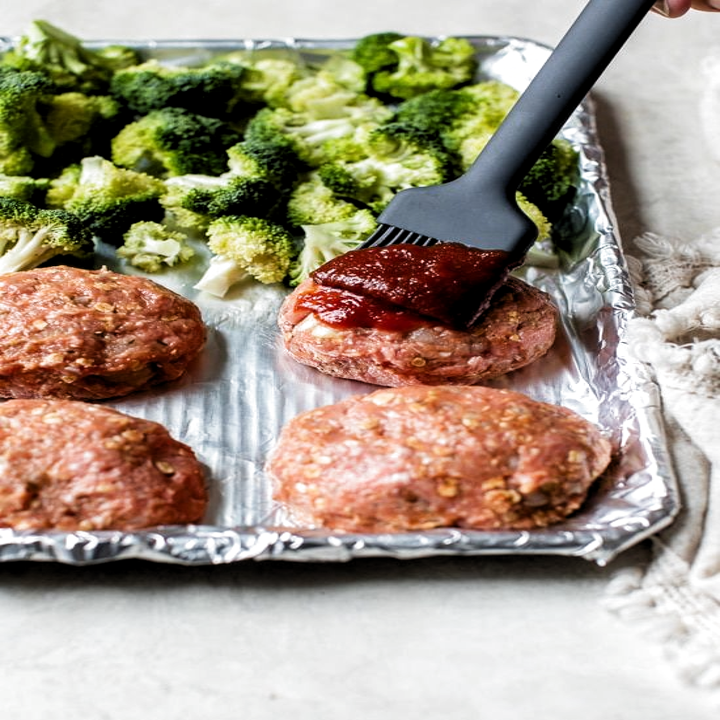 Brushing the turkey meatloafs, placed on a sheet pan with broccoli florets, with ketchup.