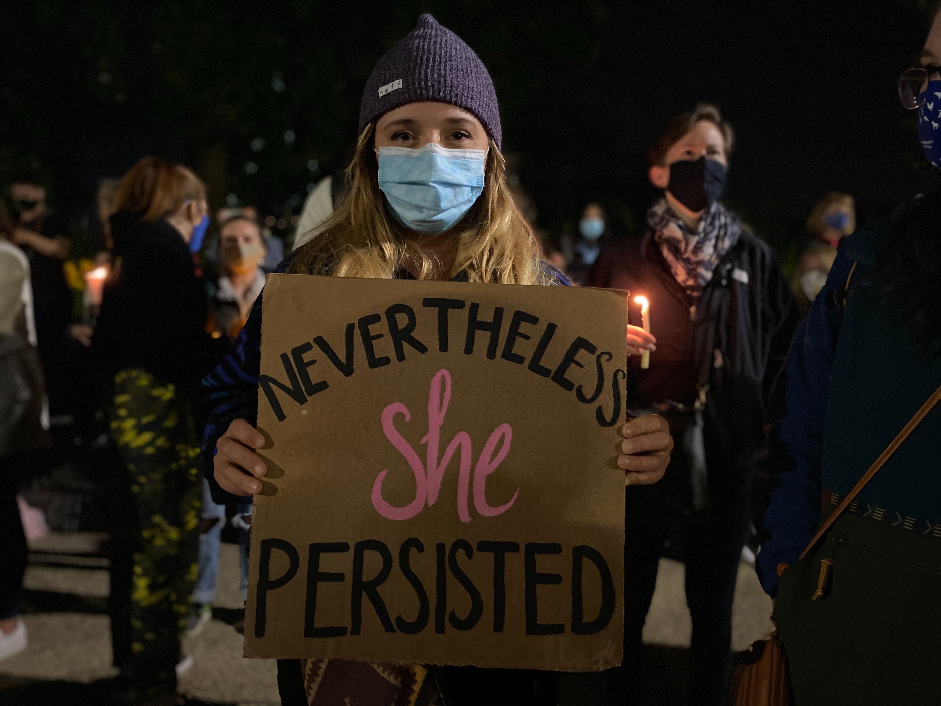 A woman wearing a beanie and a face mask holds a sign that reads &quot;Nevertheless she persisted&quot;