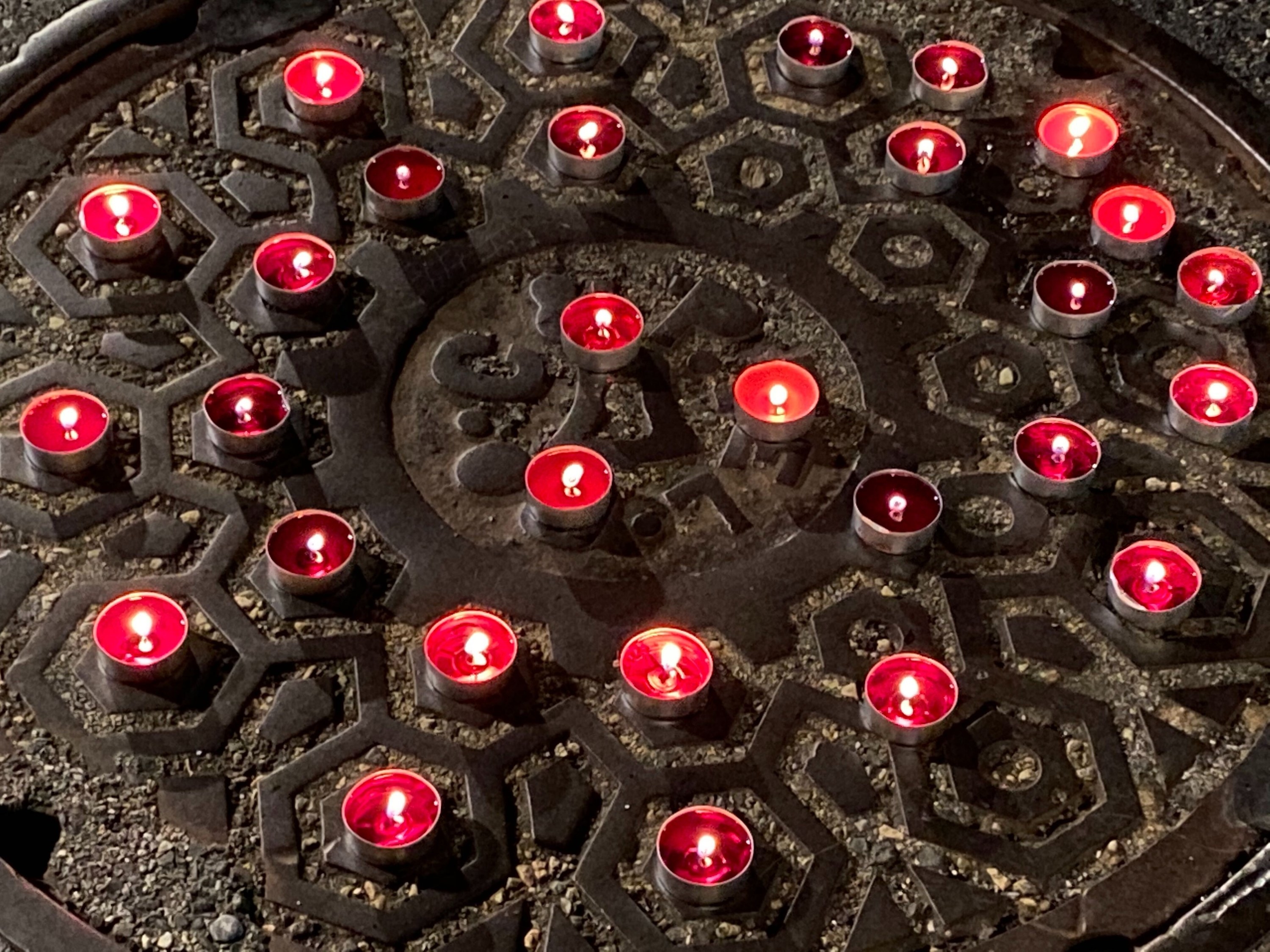 A small vigil of red candles burning atop a sewage manhole cover