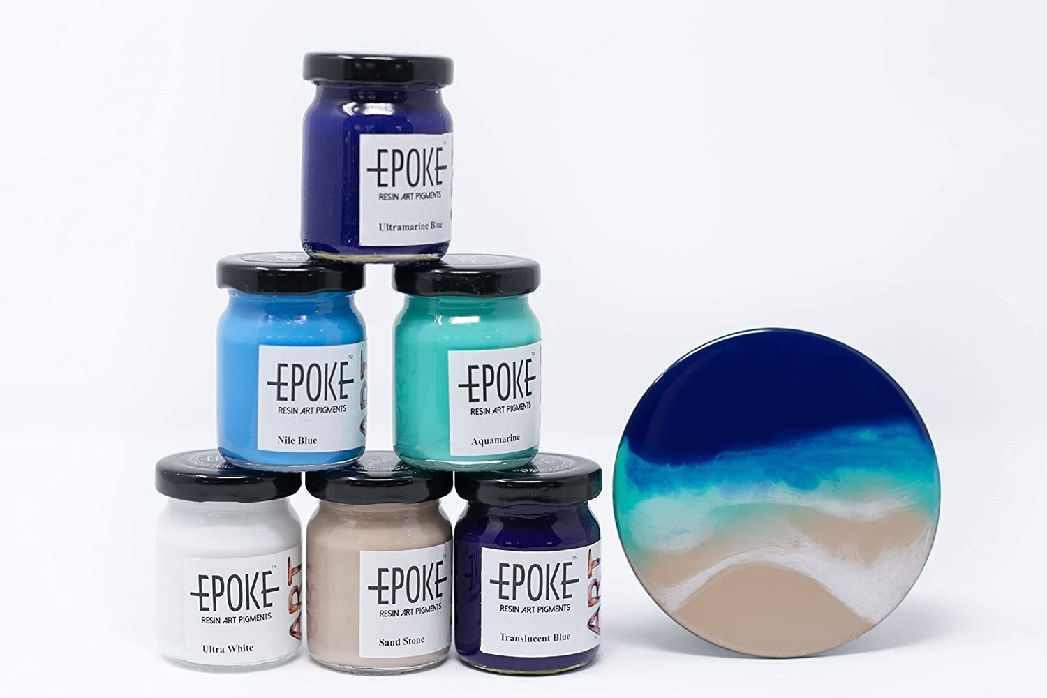 Six bottles of resin pigment in various shade of blue, sand brown and white pictured next to a resin coaster.