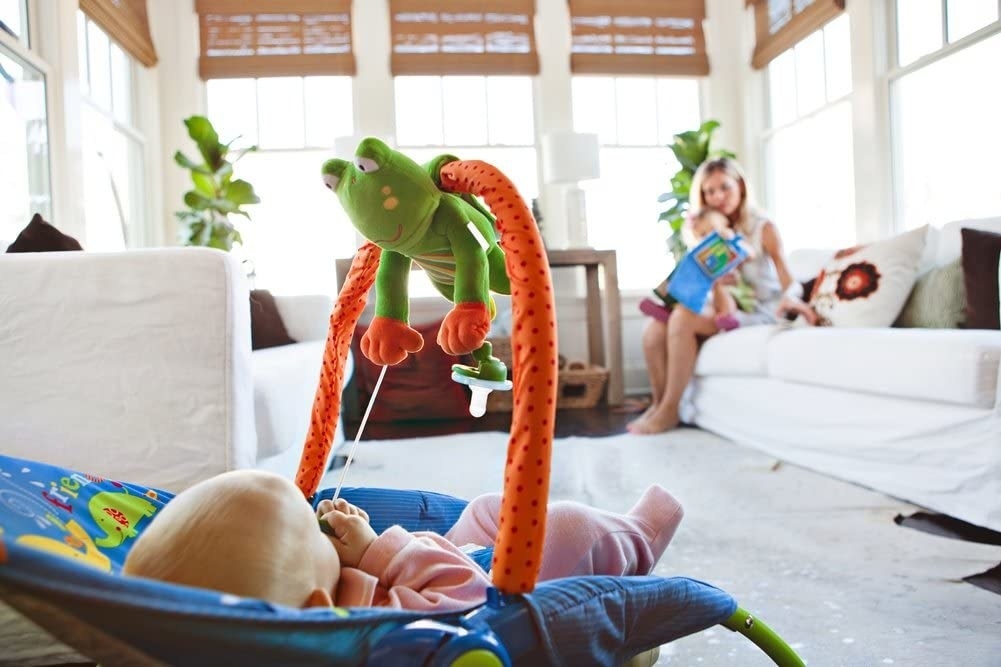 A baby sitting in a bouncer on the floor with a frog-shaped toy hanging above them