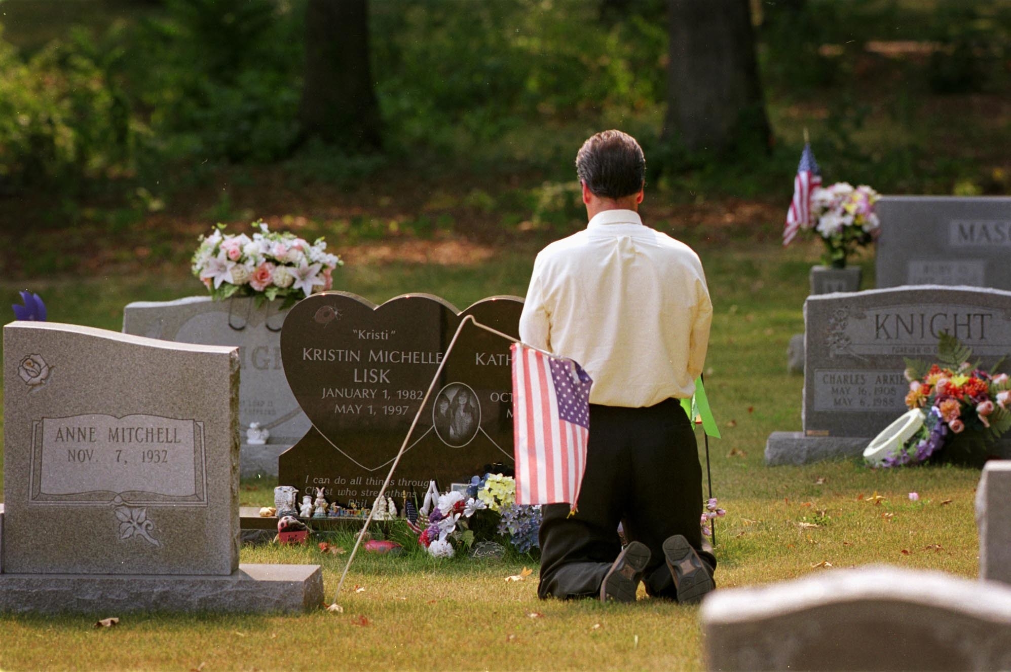 A male FBI agent in a white shirt kneels in a graveyard in front of the heart-shaped tombstone of the Lisk sisters.