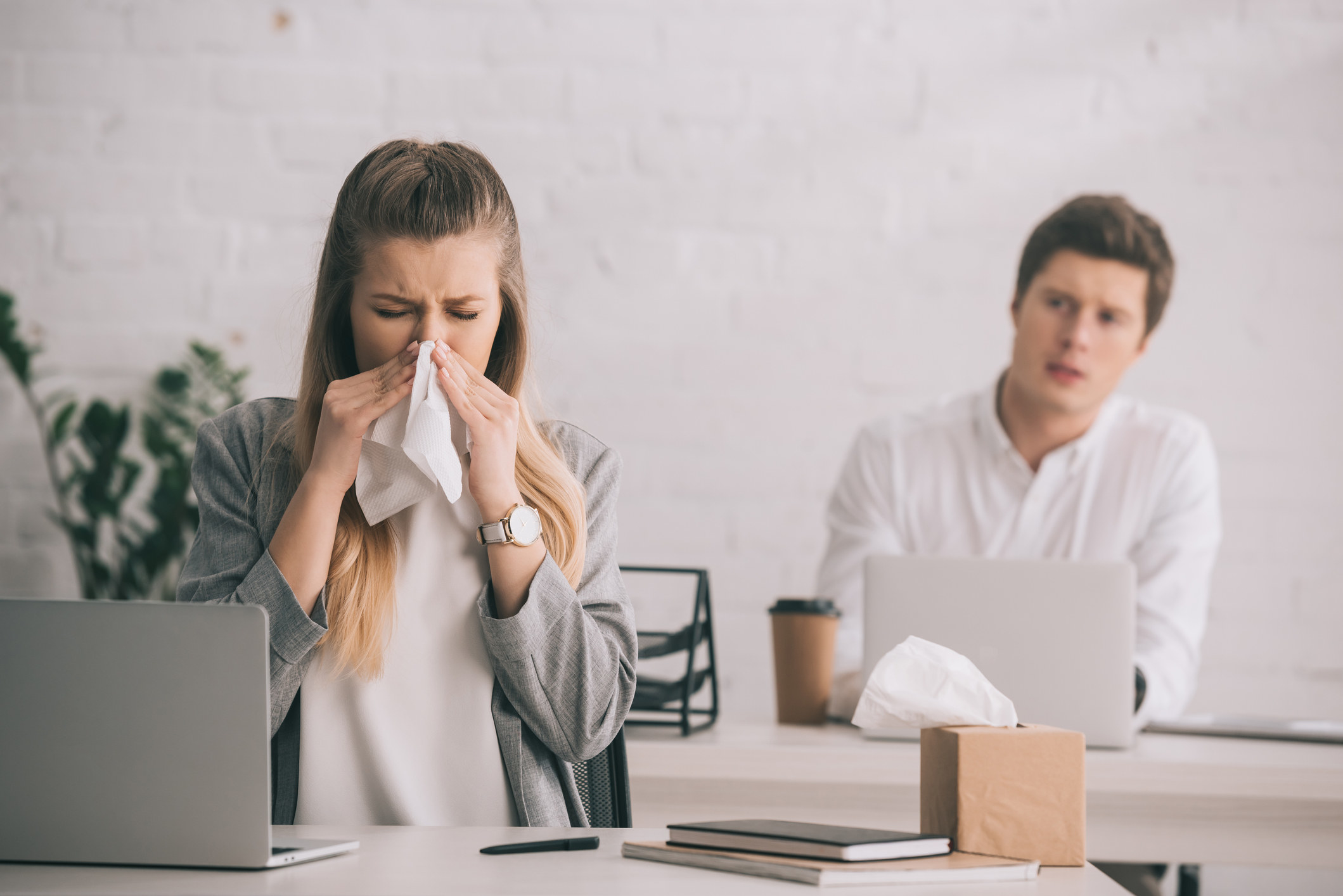 Business person sneezing into a tissue near a coworker. 