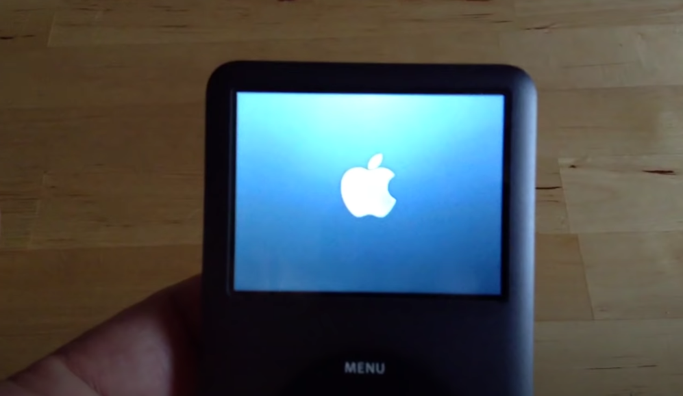 A screenshot of hand holding a black iPod Classic resetting with the Apple logo showing