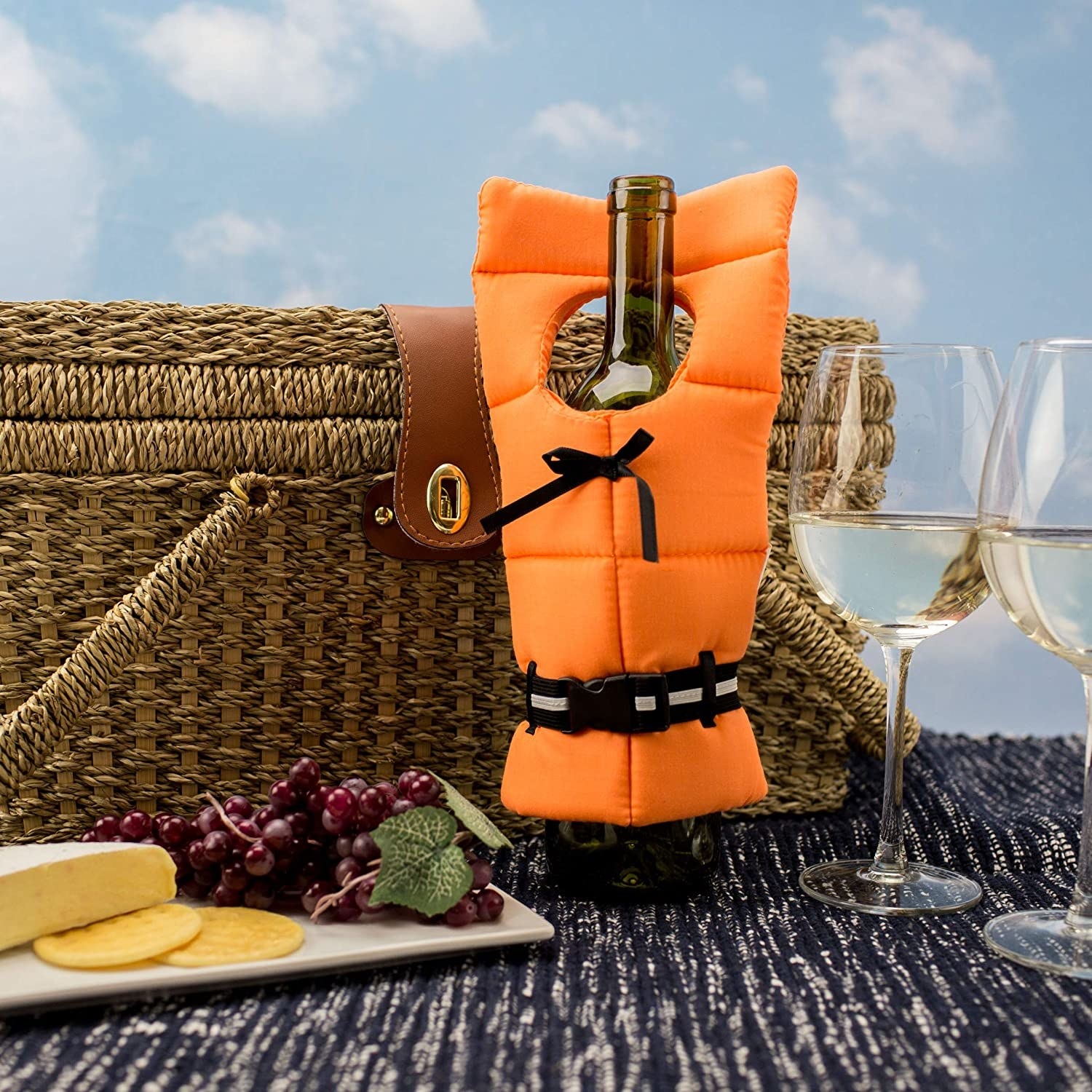 A bottle of wine with the lifejacket on it surrounded by a cheese plate, wine glasses, and picnic basket.