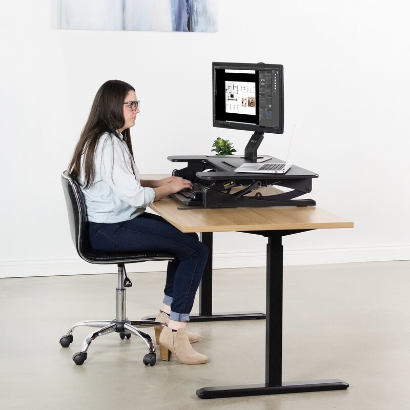 Model sitting at desk with the monitor riser holding a laptop and monitor 