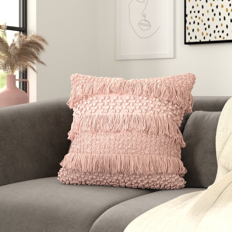 Pink throw pillow with stripes of fringe of different lengths on a couch. 