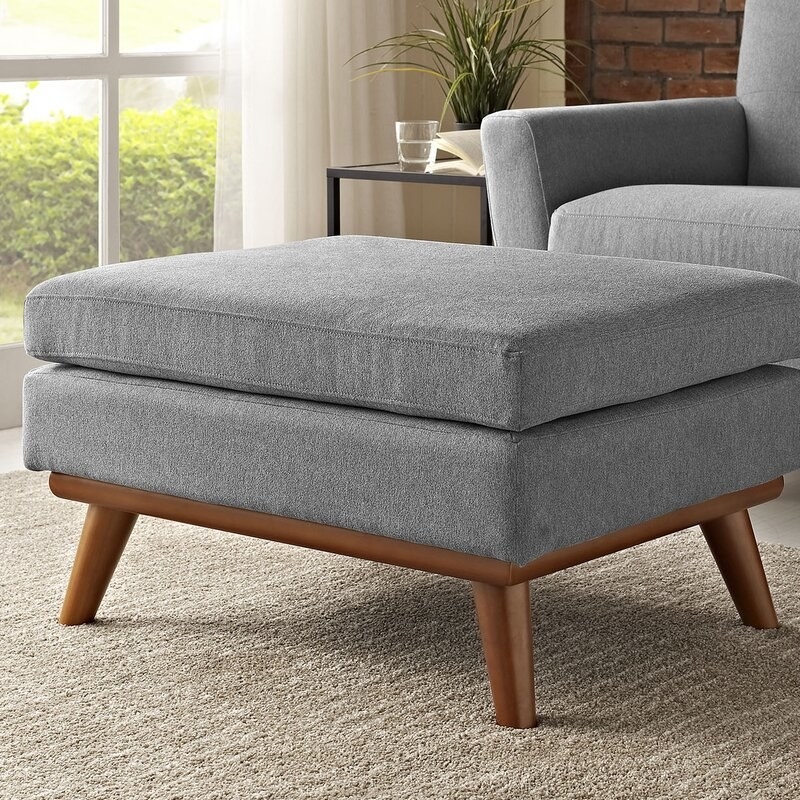Grey ottoman with a cushion on top and brown legs. 