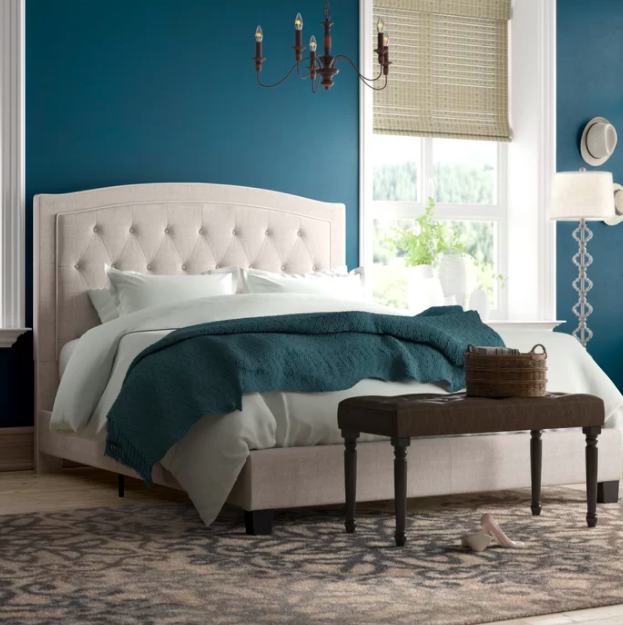 Beige upholstered bed with tufted headboard, white sheets, and a dark green throw blanket in a teal-colored bedroom