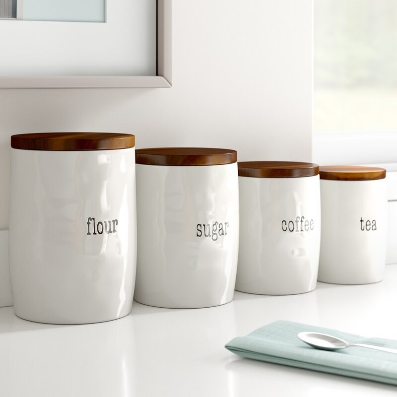 Set of four white ceramic canisters with brown lids and the words flour, sugar, coffee, tea on individual jars 