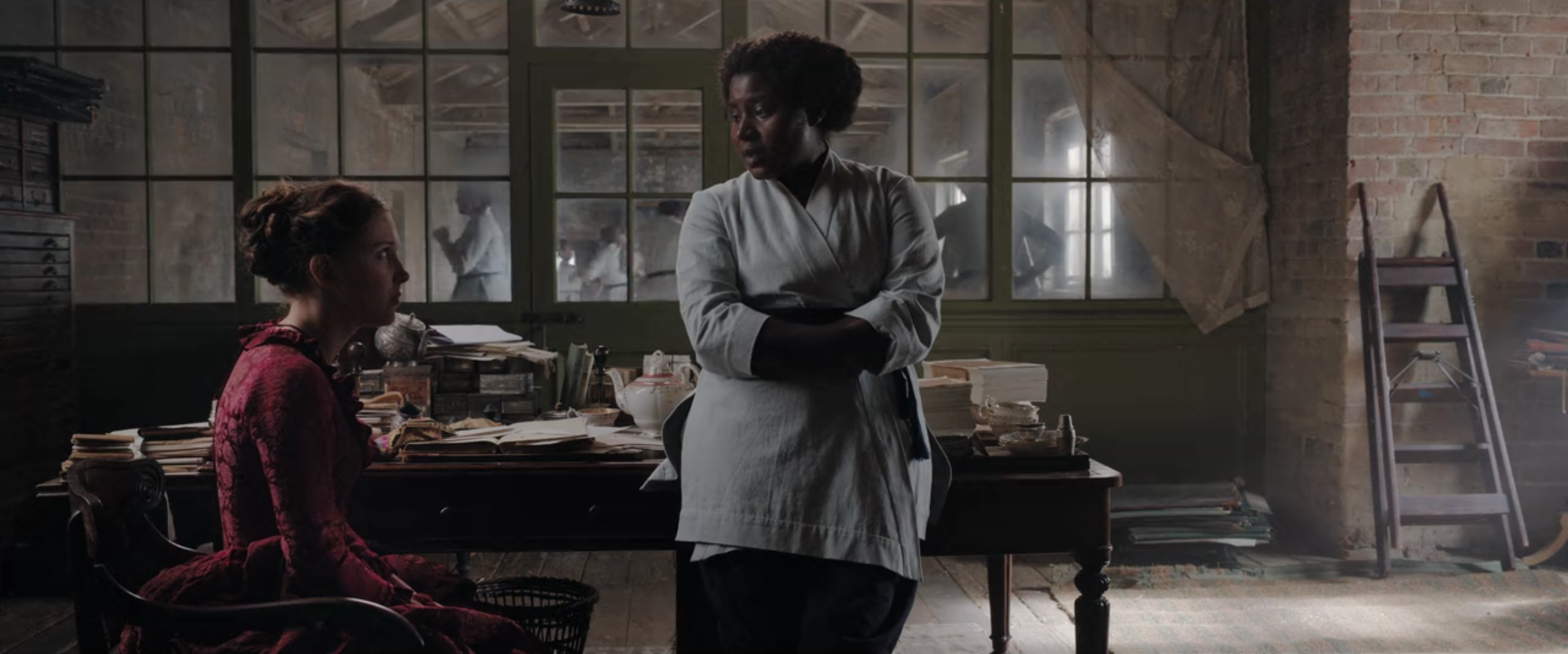 Edith leaning against her desk and talking to Enola, who is seated.