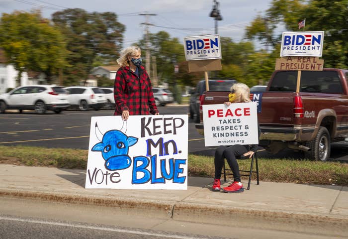 People on a sidewalk hold signs that say "Peace takes respect and kindness" and "Vote — Keep Minnesota blue"