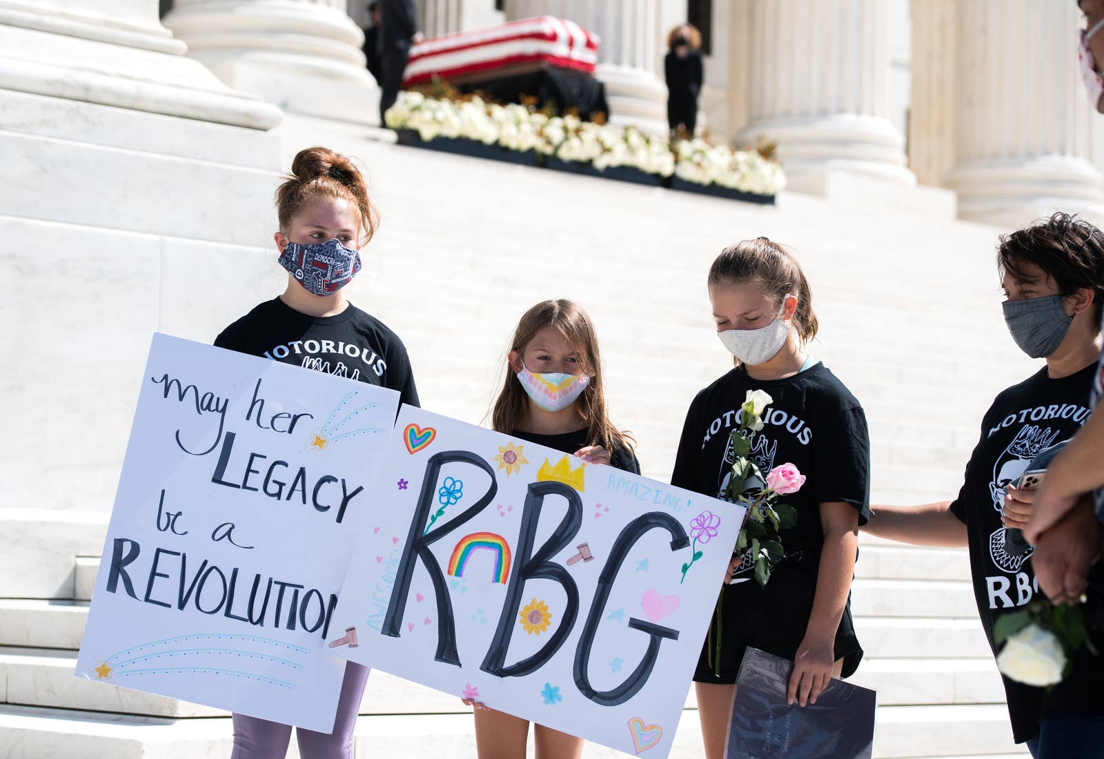Three girls carry signs saying RGB and may her legacy be a revolution
