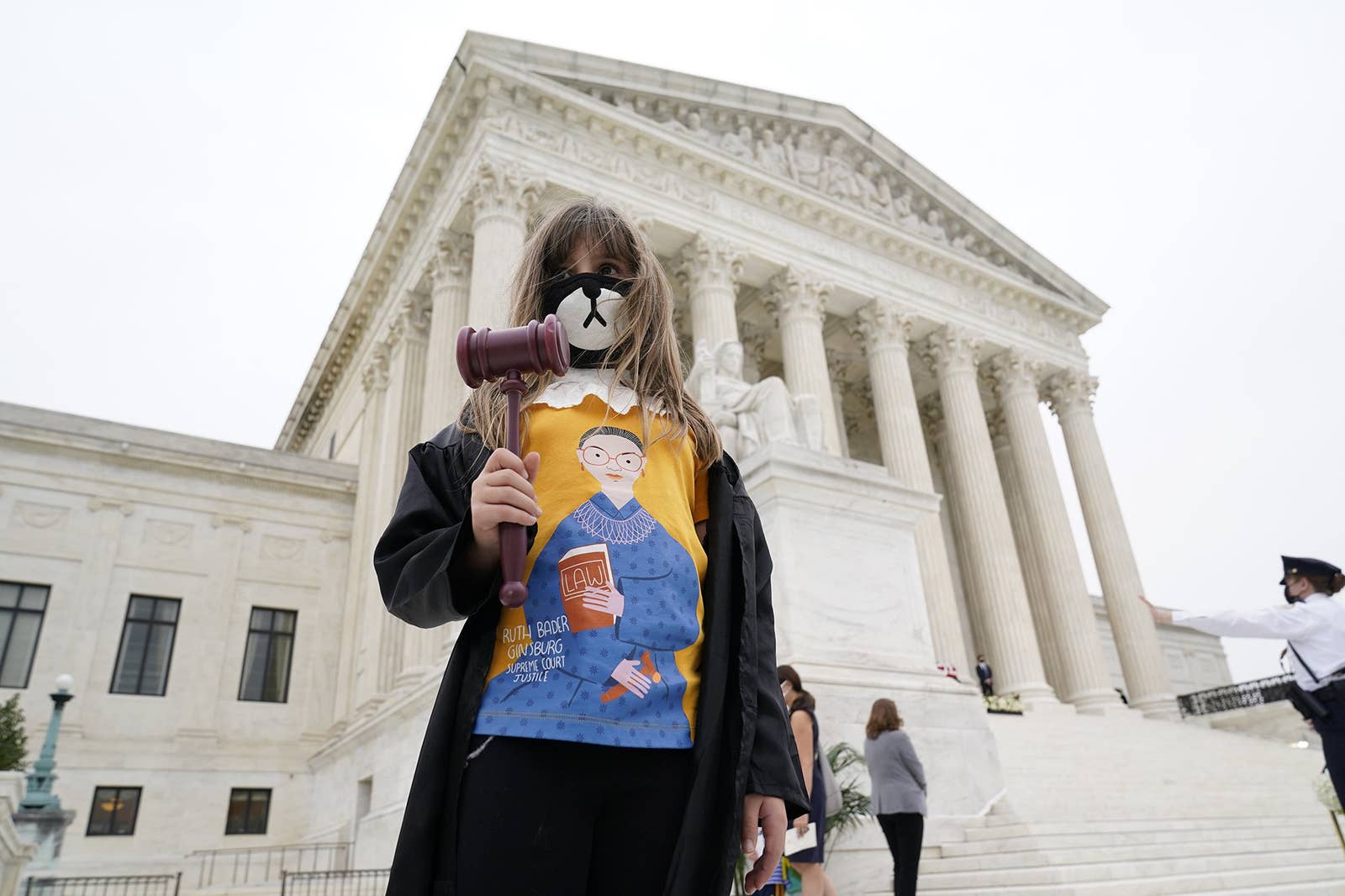 A girl wearing an RGB shirt carries a gavel in front of the Supreme Court