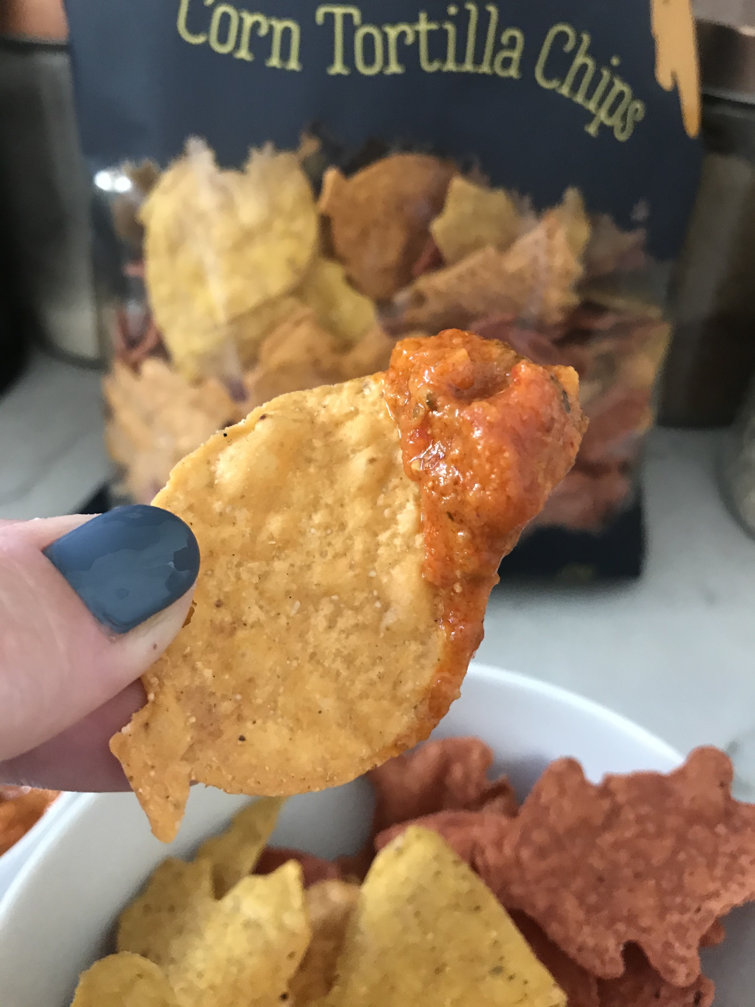 A finger holds up a leaf-shaped tortilla chip dipped in a creamy red salsa with the bag of chips in the background