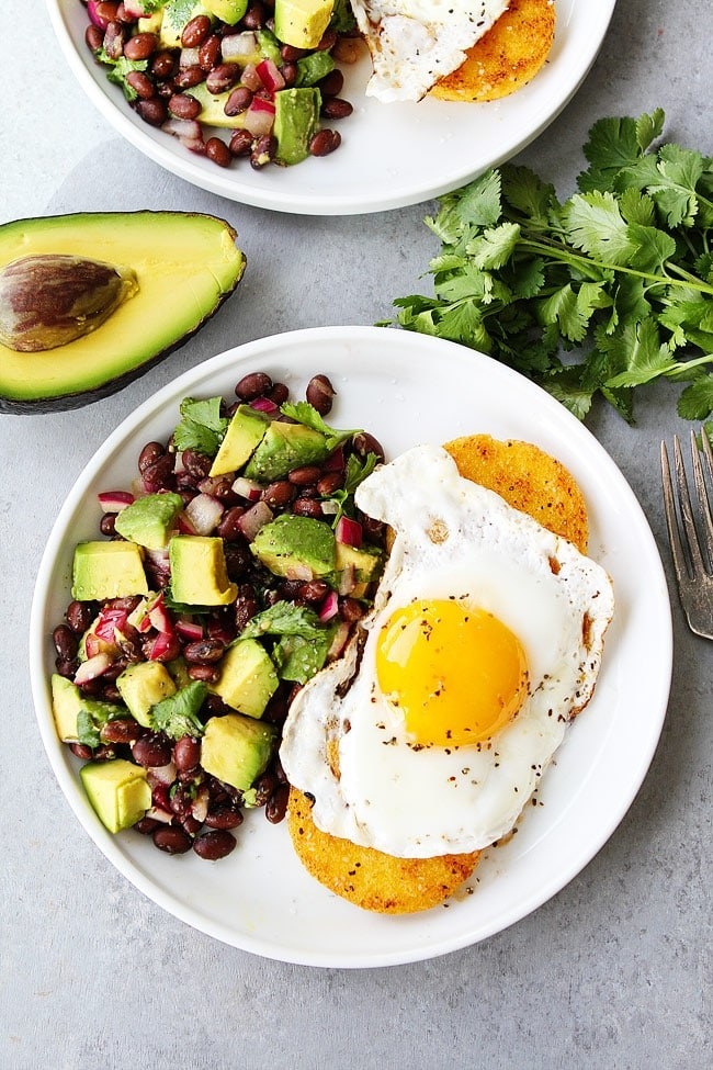 A plate with two crispy polenta cakes, each topped with a golden fried egg, with a pile of avocado and bean salsa on the side.