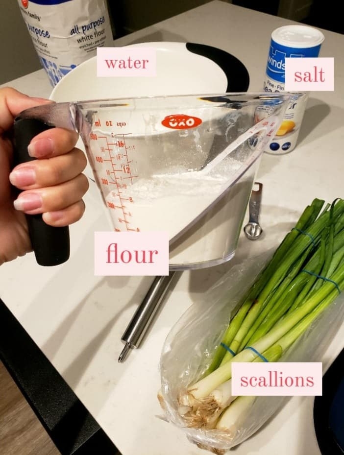 The writer holds a cup of flour over a scallions, salt, a bowl, a bag of flour, and a whisk on the counter