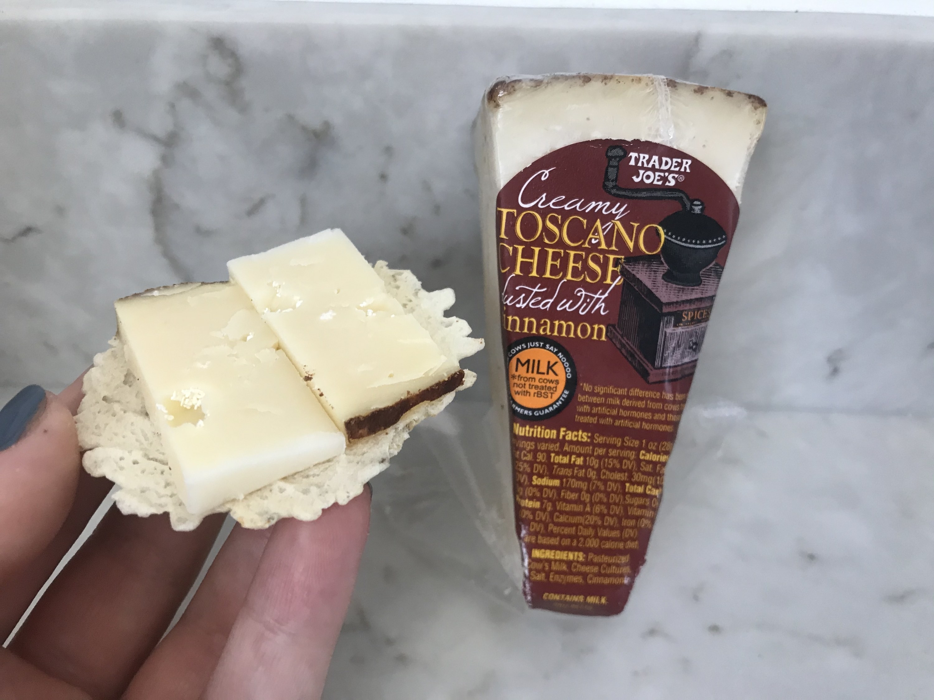 A wedge of cheese in a dark red packaging leans against a marble backdrop next to a hand holding up a cracker topped with two thin slices of cheese
