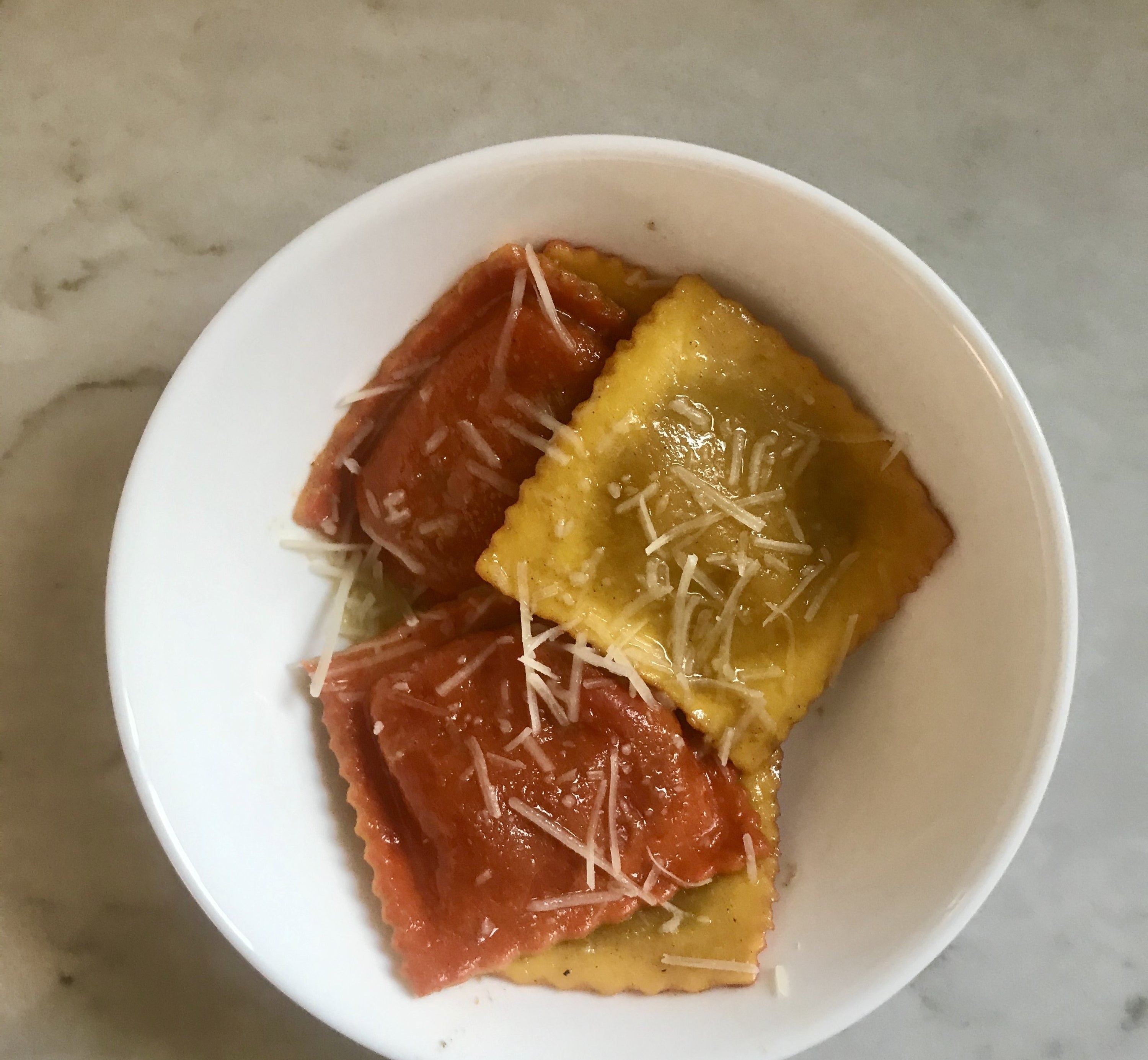 A white bowl filled with orange and yellow raviolis topped with a butter sauce and shreds of parmesan cheese